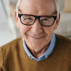 An older man wearing glasses and a dark tan sweater, smiling after receiving his All-On-4 implants